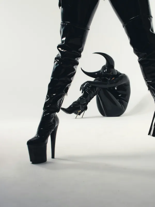 Person in Latex-Overall mit Hörnern sitzt am Boden, während eine andere Figur in Lackstiefeln dominant im Vordergrund steht.