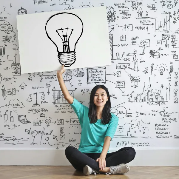 Frau mit Glühbirne auf Whiteboard vor Wand voller Ideen – Symbol für Gründung und Kreativität