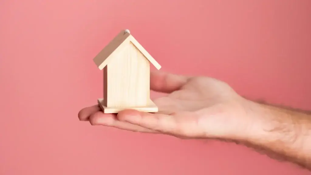 Hand hält ein kleines Holzhausmodell vor rosa Hintergrund – Symbol für die Suche nach dem passenden Swingerclub Standort