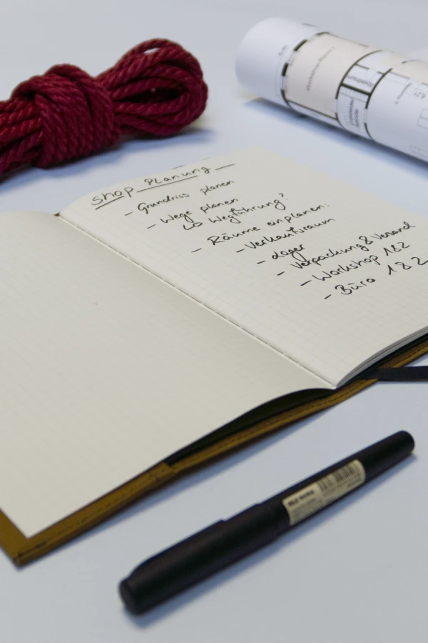 Aufgeschlagenes Notizbuch mit handgeschriebener Shop-Planung, daneben ein aufgerollter Grundriss und rotes Bondage-Seil