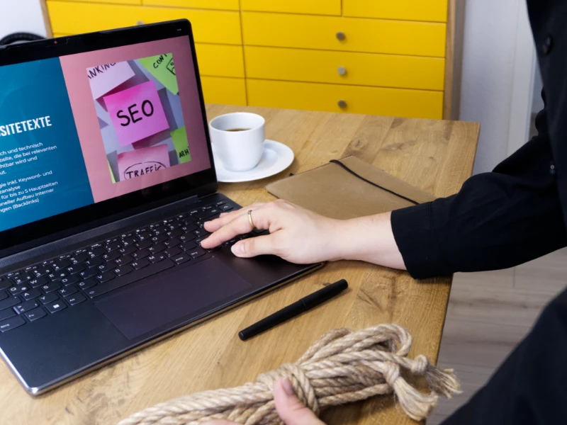 Laptop zeigt SEO-Präsentation mit Begriffen wie Websitetexte, Traffic und Ranking, daneben ein Bondage-Seil auf dem Schreibtisch